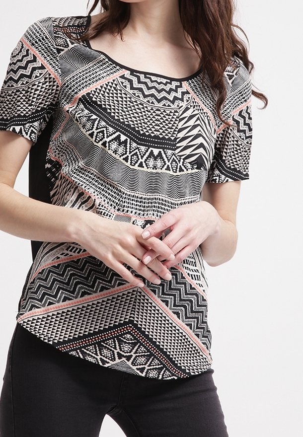 Femme portant un haut à manches courtes avec des motifs géométriques et en zigzag noirs, blancs et roses, mains croisées à la taille, pantalon noir visible.