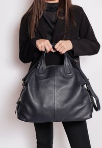 Donna in un completo nero che tiene una grande borsa in pelle nera textured con manici corti e lunghi su uno sfondo bianco.