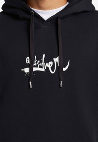 Sudadera negra con cordones, que presenta un logo blanco en un estilo de escritura en pincel. Hecha de una tela suave y texturizada.