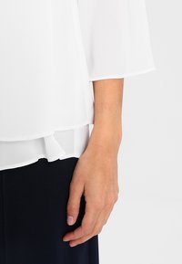 Weiße, leichte Kreppbluse mit fließenden Ärmeln und gestuftem Saum, drapiert über einem dunklen Stoff darunter, wodurch ein sanfter Texturkontrast entsteht.