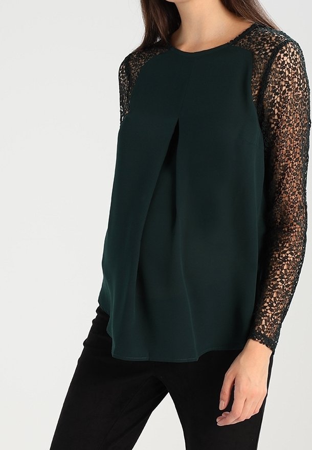 Blouse vert foncé avec des manches en dentelle, présentant un col rond et un devant plissé. Tissu lisse avec des accents en dentelle texturée.