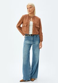 Bruine Suède cropped jas met twee voorkeurszakken, drukknoopsluiting en een ronde halslijn, gecombineerd met lichtblauwe wijde jeans en bruine schoenen.