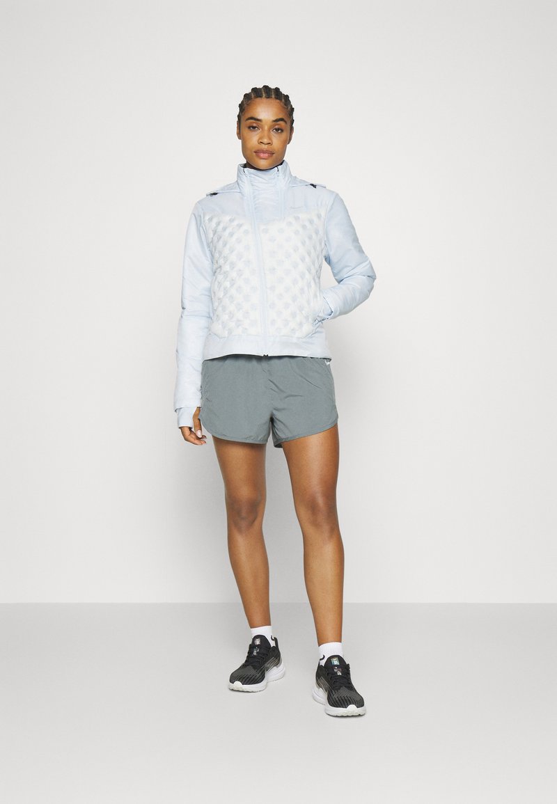 Frau in einer hellblauen Steppjacke, grauen Sportshorts, weißen Socken und schwarzen Laufschuhen, die vor einem schlichten Hintergrund steht.