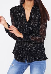 Schwarze durchsichtige Bluse mit weißen Punkten, V-Ausschnitt, langen Ärmeln und einem Saum, der an den Seiten leicht nach außen geschwungen ist. Kombiniert mit ausgebleichten Jeans.