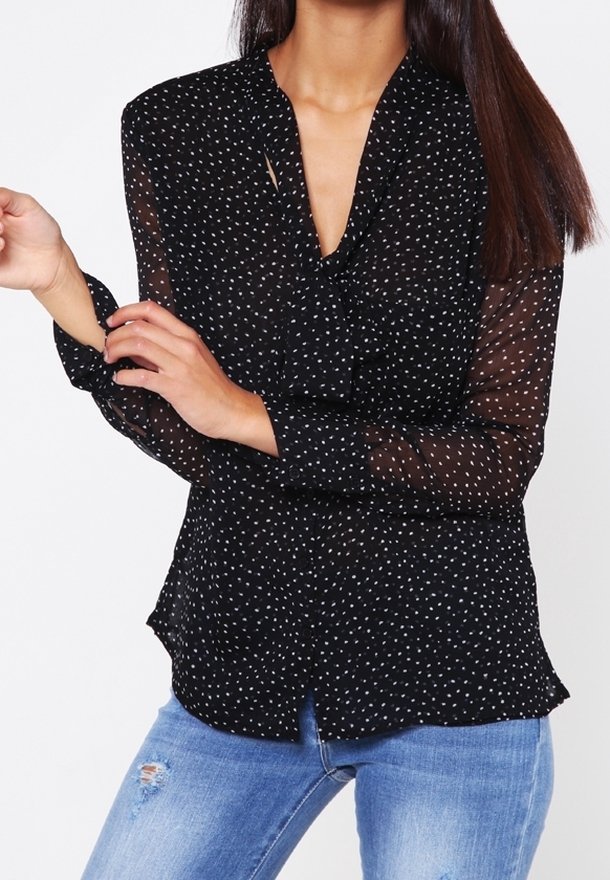 Schwarze durchsichtige Bluse mit weißen Punkten, V-Ausschnitt, langen Ärmeln und einem Saum, der an den Seiten leicht nach außen geschwungen ist. Kombiniert mit ausgebleichten Jeans.