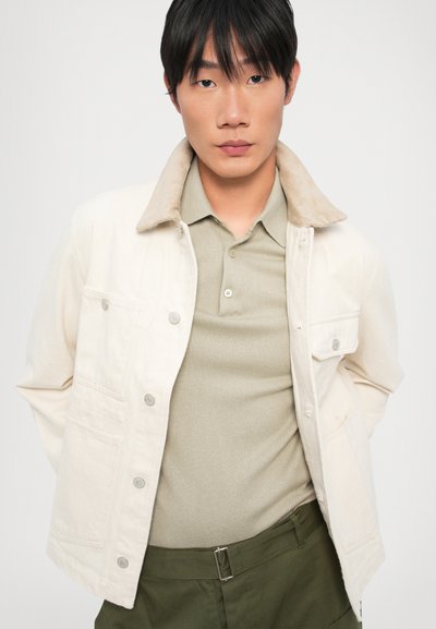 Κρεμ τζάκετ με κολάρο από καστόρι, με μπροστινές τσέπες και ασημένια κουμπιά, φορεμένο πάνω από ένα ανοιχτό πράσινο πόλο μπλουζάκι.