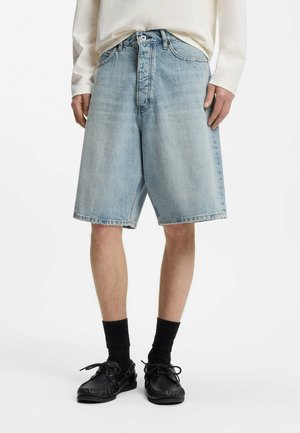 Person iført lyseblå knælange denimshorts, hvid langærmet skjorte, sorte sokker og sorte sko, stående mod hvid baggrund.