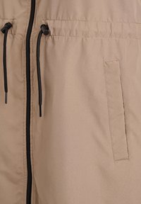Beige Windbreaker mit strukturierter Oberfläche, durchgehendem Reißverschluss, verstellbarer Kapuze mit Kordelzug und Seitentasche ohne sichtbare Verschlüsse.