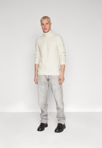 Maglione a collo alto crema con una trama diagonale, jeans grigio chiaro e scarpe nere stringate, in piedi di fronte a uno sfondo grigio uniforme.