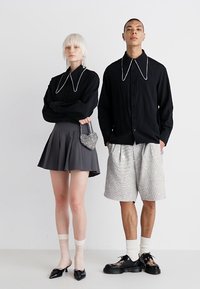 Camisa preta com botões e detalhe na gola branca, saia pregueada cinza, bolsa brilhante e calções texturizados com detalhes brancos. Sapatos e meias pretas.