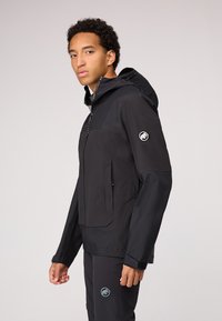 Chaqueta negra, impermeable, con capucha, paneles texturizados, cierre delantero con cremallera y bolsillos laterales. Parche de logo en la manga izquierda. Diseño ajustado.