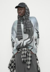 Υπερμεγέθη γκρι πουλόβερ με αφηρημένο μοτίβο, μαύρες λεπτομέρειες στα μανίκια, πολυεπίπεδη γκρι ριγωτή εσάρπα, μαύρο καπέλο και πουκάμισο με καρό σχέδιο δεμένο στη μέση.