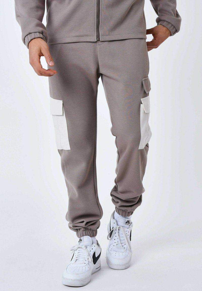 Grijze cargo broek met elastische boorden, twee zijzakken en witte accenten. Het materiaal lijkt zacht en rekbaar, geschikt voor casual wear.