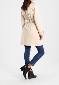 Kobieta w beżowym płaszczu z paskiem, niebieskich wąskich dżinsach z rozcięciami na kostkach i czarnych botkach na obcasie, stojąca na jednolitym jasnym tle.
