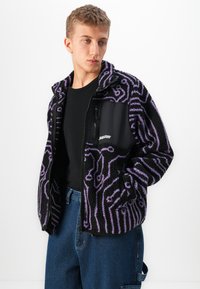 Jauns vīrietis melnā un purpursarkanā ar rakstu flīsa jakā, melnā T-kreklā un zilās džinsās, pozē ar rokām jakas kabatās uz baltā fona.