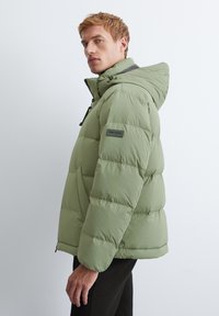 Doudoune verte clair avec capuche, dotée de sections matelassées et d'une fermeture éclair à l'avant. Comprend un patch logo sur le bras gauche.