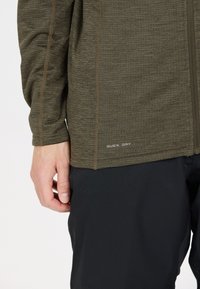 Olivengrøn hurtig-tørrende jakke med tekstureret stof, kontrasterende sømme og lynlås, vist med sorte bukser.