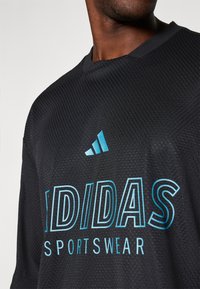 Melnā sporta kreklā ar strukturētu audumu, ar gaiši zilu "adidas" logo un "SPORTA APĢĒRBS" tekstu, apaļā apkaklītē un īsajām piedurknēm.