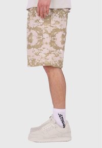 Beige camouflage shorts med tekstureret stof, knælængde, med en lodret søm og et lille hvidt logo. Parret med hvide sneakers og sokker.