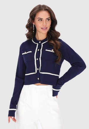 Vrouw draagt een marineblauw cropped vest met witte bies en parelknopen, gecombineerd met een witte broek met hoge taille tegen een effen achtergrond.