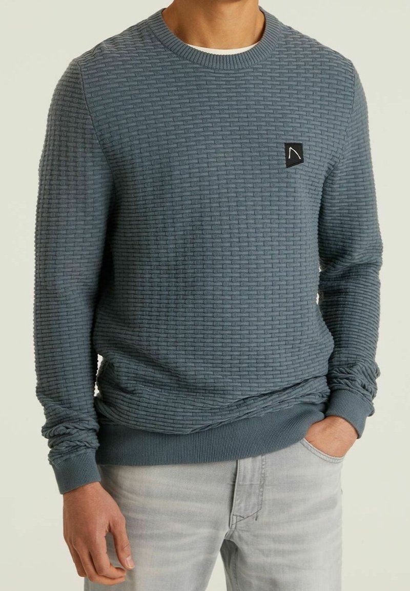 Strukturierter blauer Sweatshirt mit geripptem Ausschnitt und Bündchen, mit einem kleinen schwarzen Logopatch. Getragen über hellgrauen Jeans.
