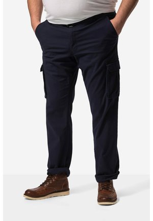 Man wearing navy cargo pants, brown lace-up boots, and a white shirt with hands in pockets, standing against plain background.