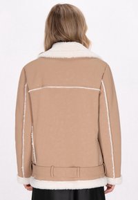 Frau, die eine beigefarbene Lammfelljacke mit weißem, flauschigem Futter und Gürteldetails trägt, von hinten vor einem einfarbigen weißen Hintergrund gezeigt.