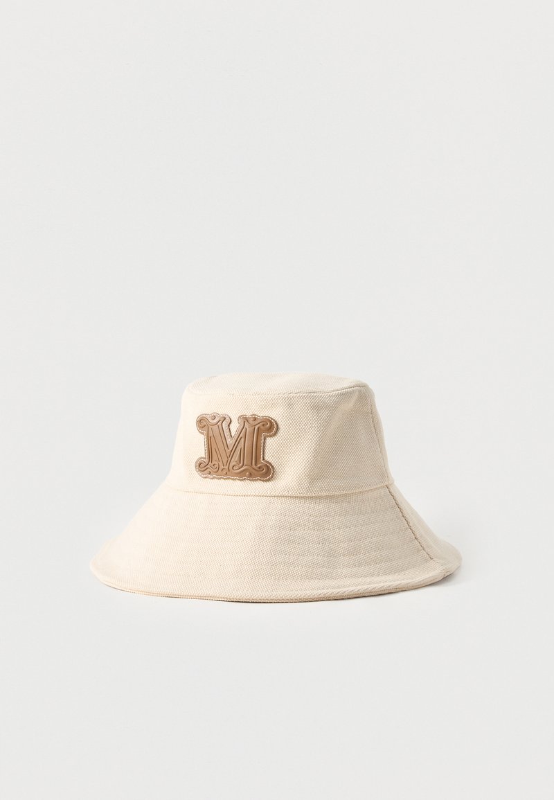 Μπεζ υφασμάτινο καπέλο τύπου bucket με φαρδύ γείσο και ανάγλυφο καφέ μπατς με το γράμμα "M" στο κέντρο μπροστά.