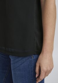 Bras droit d'une personne portant un haut noir et un jean bleu sur un fond uni.