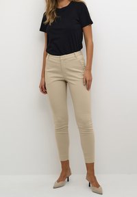 Pantaloni beige su misura con una vestibilità slim, caratterizzati da una vita piatta, passanti per cintura e tasche laterali, abbinati a una maglietta nera e tacchi beige.