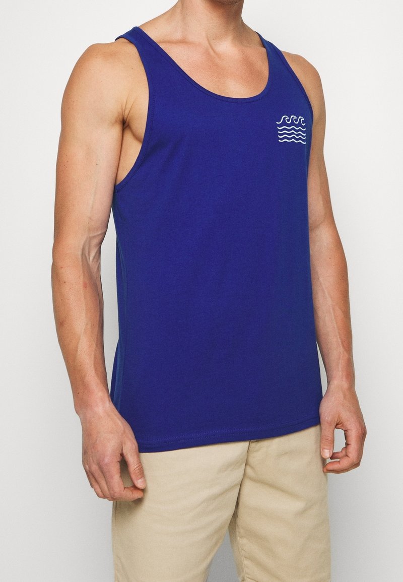 Débardeur en coton bleu avec un col rond et un petit motif de vagues blanches sur le côté gauche de la poitrine. Caractérisé par une coupe décontractée et des coutures latérales.