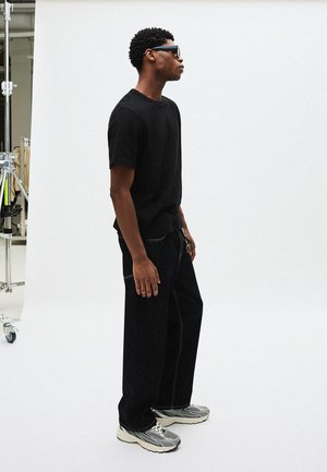 Crna majica kratkih rukava, opuštenog kroja; tamne traperice s kontrastnim bijelim šavovima; sive tenisice s teksturiranim uzorkom; sunčane naočale.