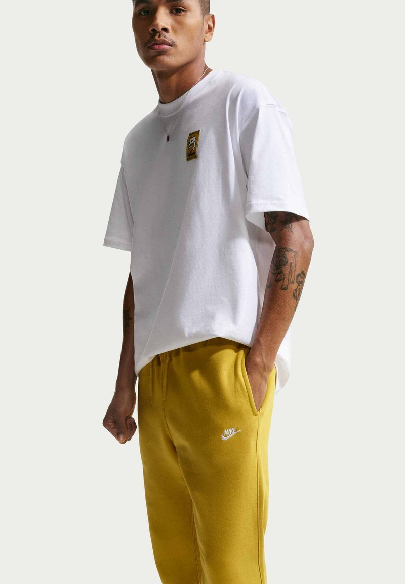 Homme portant un T-shirt blanc avec un petit motif sur la poitrine et un pantalon de survêtement Nike jaune moutarde, debout avec une main dans la poche.