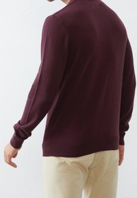 Suéter de punto burdeos con mangas largas, puños acanalados y dobladillo. Lleva puesto sobre pantalones de color beige claro, mostrando una textura suave.