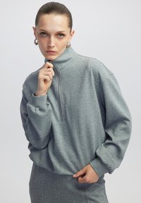 Grijze rits-hoodie van zacht materiaal, met een hoge kraag, lange mouwen en een ontspannen pasvorm, met zichtbare stikseldetails.
