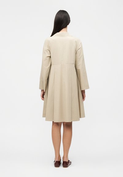 Γυναίκα με μακριά σκουρόχρωμα μαλλιά, φορώντας ένα μπεζ παλτό μήκους μέχρι το γόνατο με μακριά μανίκια, που στέκεται γυρισμένη από πίσω και καφέ επίπεδα παπούτσια.