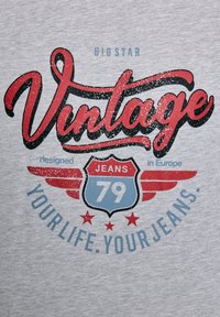 Pilkas marškinėliai su raudonais ir mėlynais retro stiliaus užrašais. Pavaizduota skydelio dizainas su "JEANS 79" ir sparnais, nusidėvėjęs tekstūros efektas bei žvaigždžių akcentai.