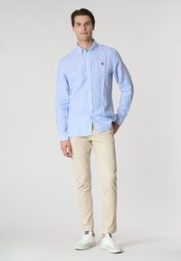 Camicia a righe blu chiaro con bottoni, pantaloni beige e sneakers bianche. La camicia presenta un piccolo logo e un colletto classico. Texture liscia.
