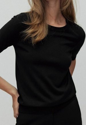 Kvinde med lysebrunt hår iført en enkel sort kortærmet skjorte og sorte bukser, hånden hvilende på hoften, neutral baggrund.