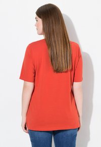 T-shirt rossa in cotone con maniche corte, vestibilità rilassata e scollatura rotonda, abbinata a jeans blu, vista da dietro su uno sfondo bianco.