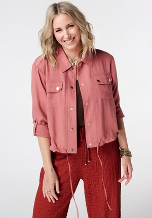 Femme souriante aux cheveux blonds, portant une veste boutonnée rose poudré et un pantalon rouge texturé, accessoirisée de bijoux et bagues en or.