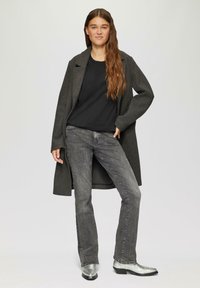 Lange grijze jas over een zwart shirt, gecombineerd met grijze jeans en zilveren enkellaarsjes. Zachte textuur, relaxte pasvorm en minimalistisch ontwerp.