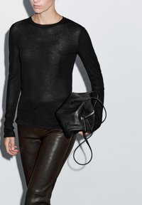 Person iført en gennemsigtig sort langærmet top og mørke læderbukser, der holder en sort læderhåndtaske med lynlås og rem.
