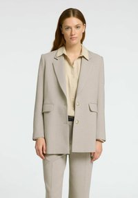 Blazer ajustado de color beige claro con corte estructurado, solapas de muesca, dos bolsillos frontales y cierre de un solo botón, combinado con una camisa crema.