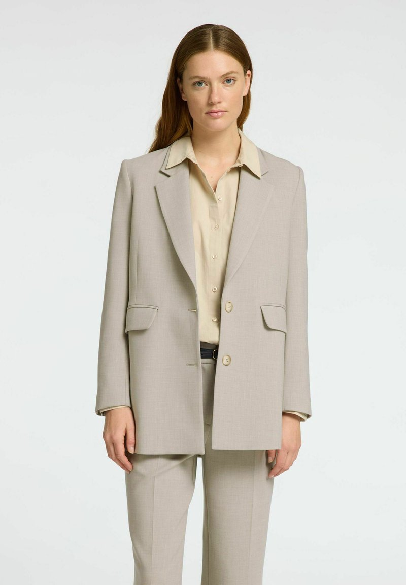 Blazer ajustado de color beige claro con corte estructurado, solapas de muesca, dos bolsillos frontales y cierre de un solo botón, combinado con una camisa crema.