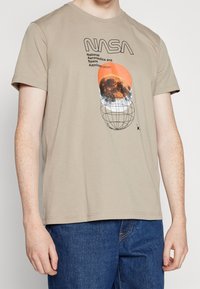 Beige bomulds-T-shirt med NASA-grafik, orange cirkulært accent, sort tekst og globusdesign. Korte ærmer og rund halsudskæring.