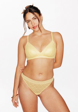 Donna con capelli castani raccolti, che indossa un reggiseno giallo di pizzo e mutandine coordinate a vita alta, posando con una mano dietro la schiena.