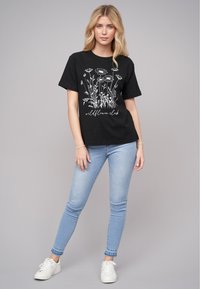 T-shirt en coton noir avec un design graphique floral blanc et le texte "wildflower club". Associé à un jean skinny bleu clair et des baskets blanches.