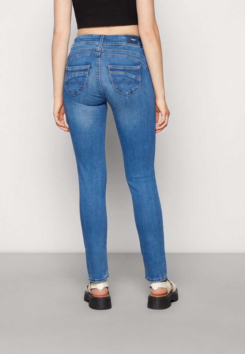 Kvinna iklädd ljusblå tajta jeans och vita platåsandaler, stående mot en enkel vit bakgrund.