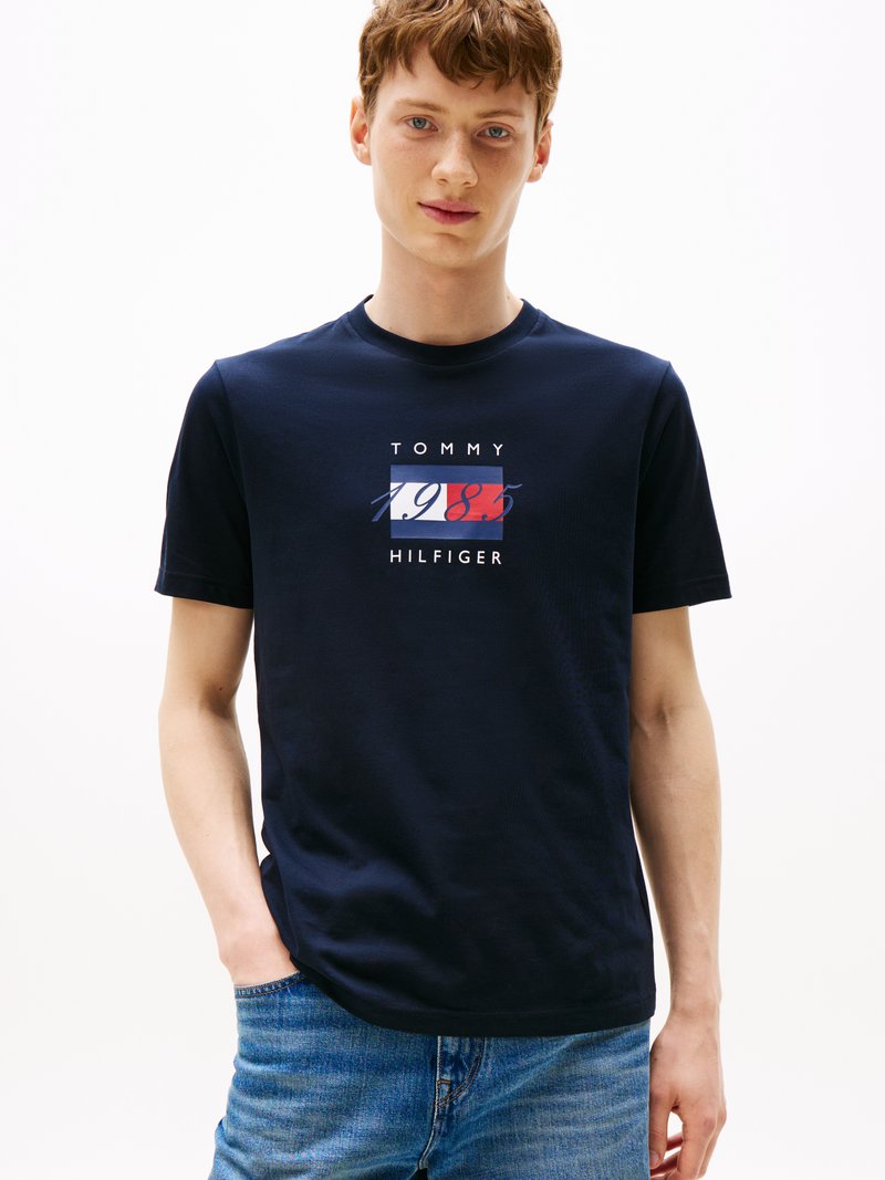 Tengerészkék pamut póló, fehér és piros grafikával, a "TOMMY 1985 HILFIGER" felirattal téglalap alakban elrendezve.